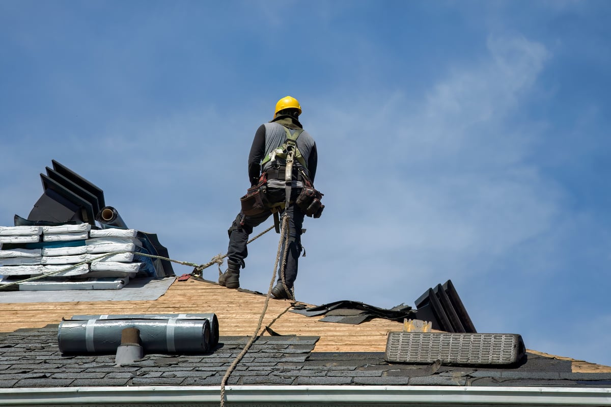 Roofing inspector examines roof shingles while crouching on residential roof at sunset with houses and trees visible below