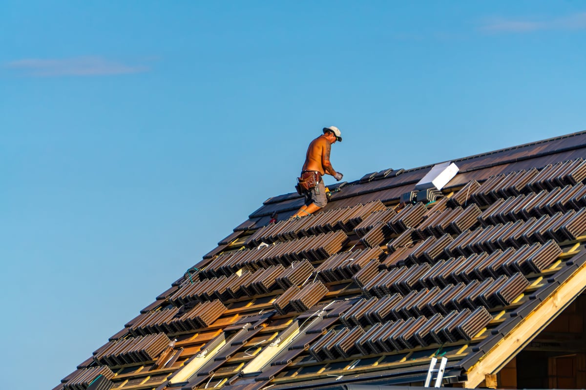 Roofing inspector examines roof shingles while crouching on residential roof at sunset with houses and trees visible below