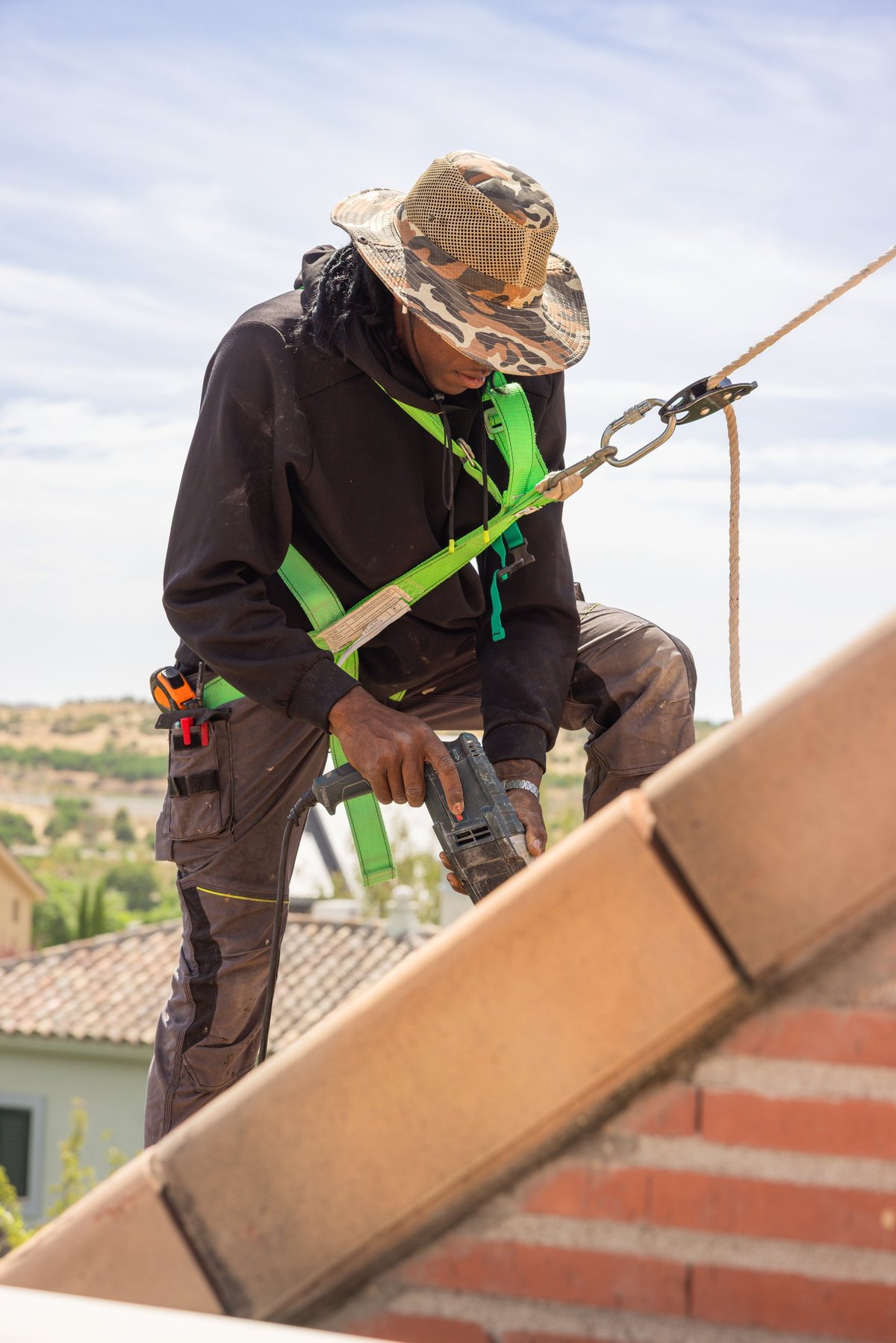 Roofing inspector examines roof shingles while crouching on residential roof at sunset with houses and trees visible below