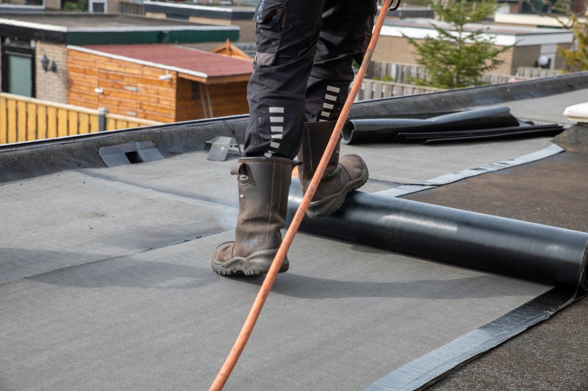 Roofing inspector examines roof shingles while crouching on residential roof at sunset with houses and trees visible below