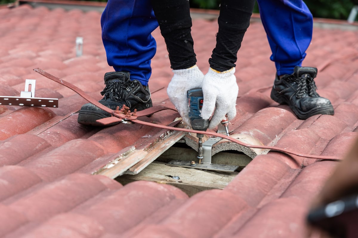 Roofing inspector examines roof shingles while crouching on residential roof at sunset with houses and trees visible below