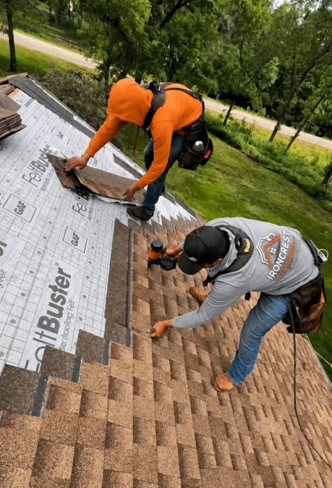 Two roofers installing asphalt shingles on a residential roof with trees visible in the background