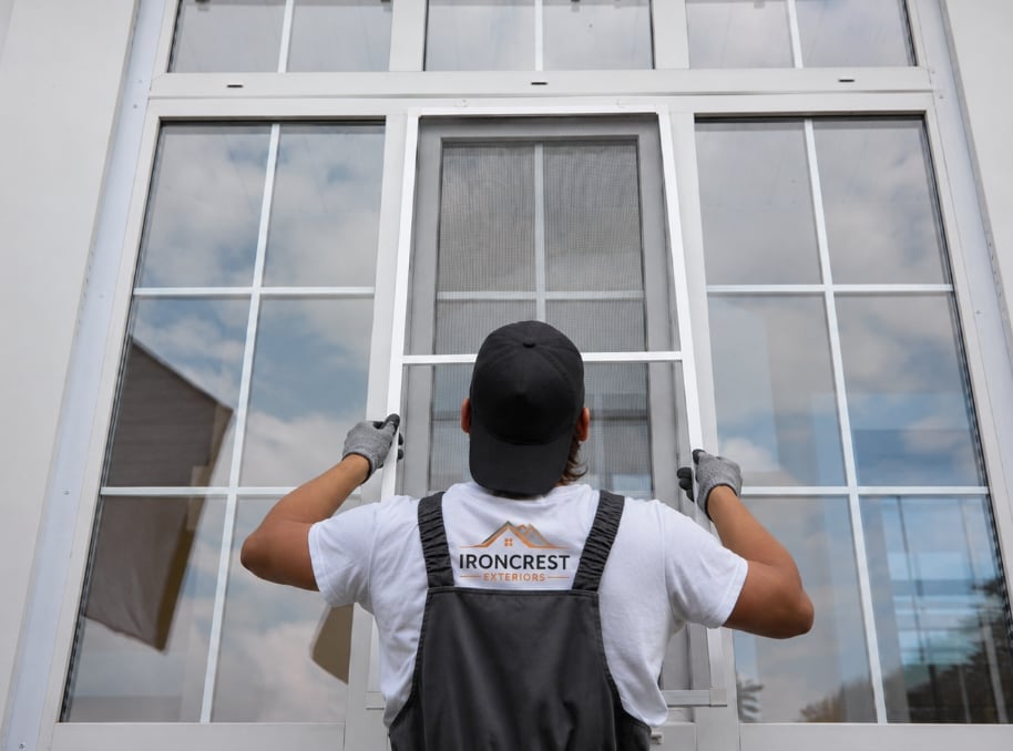 Worker in gray apron installing or repairing a large white-framed multi-pane window from outside