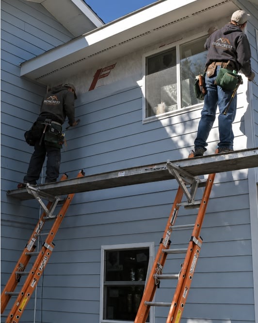 Two workers on scaffolding with orange ladders painting the exterior of a light blue residential house.