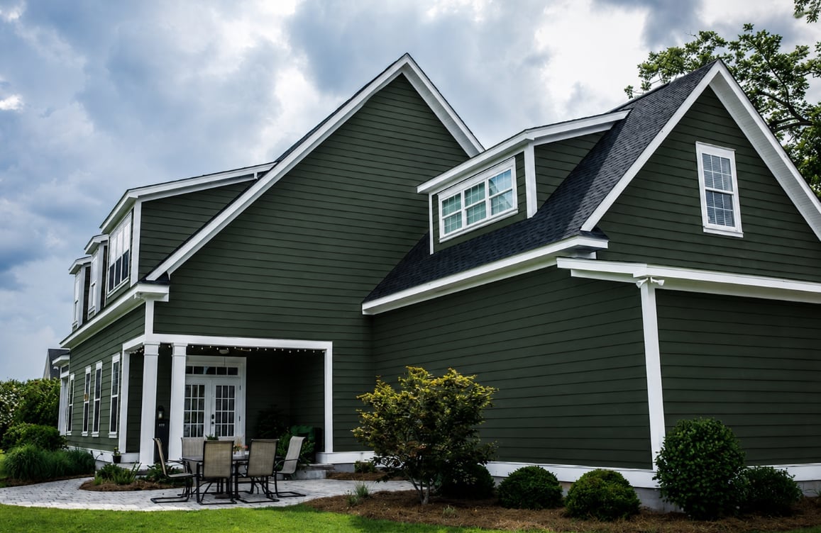 Modern two-story green house with white trim, attached garage, covered porch, and landscaped front yard under cloudy sky